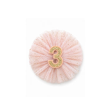 Clip circular de tul rosa con número tres dorado.