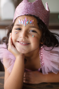 Niña sonriendo con corona rosa y cristales adhesivos en la cara apoyada en una mano