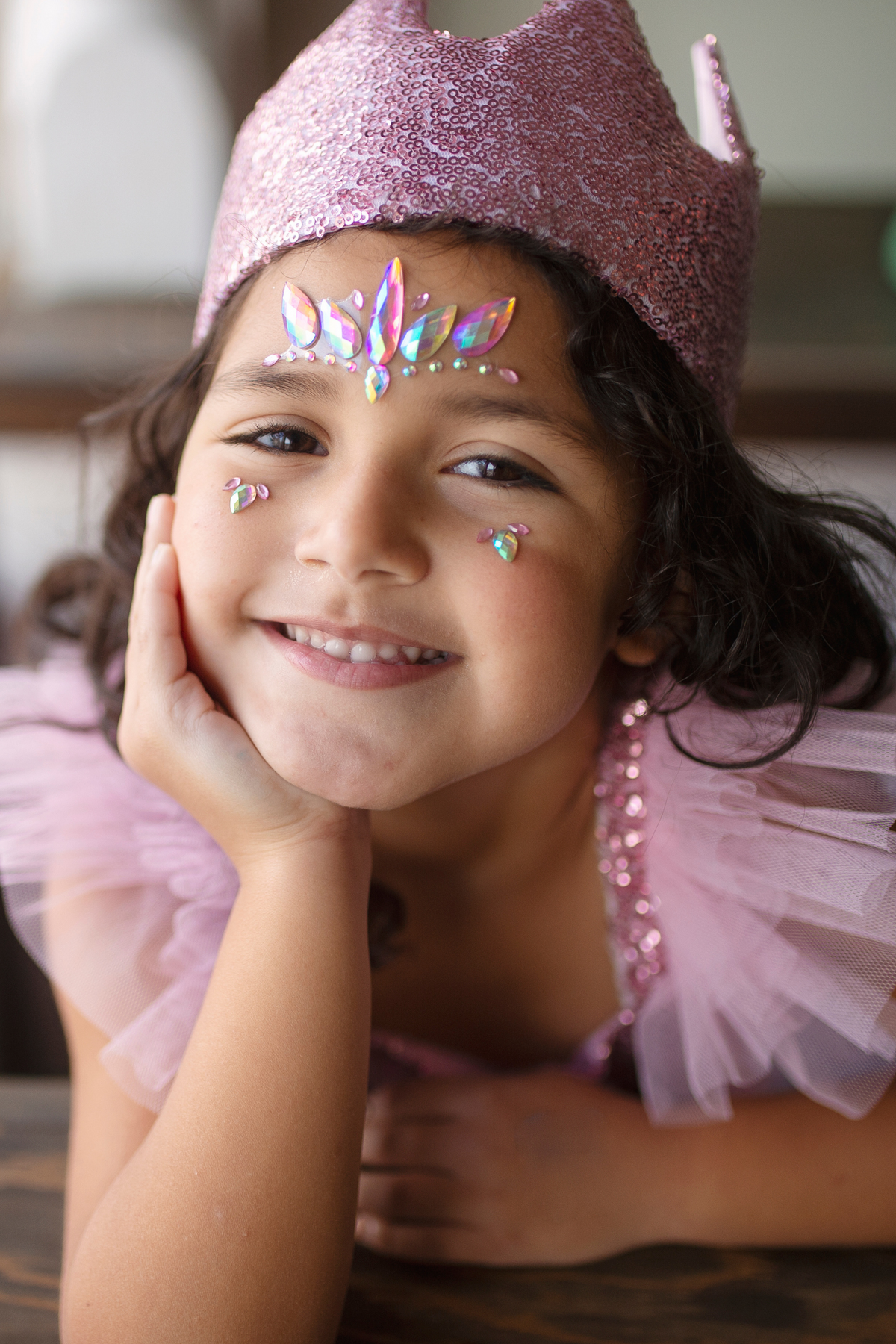 Niña sonriendo con corona rosa y cristales adhesivos en la cara apoyada en una mano